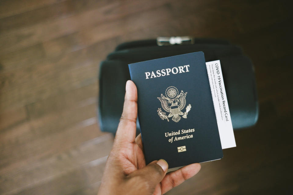 A hand holding a U.S. passport and a COVID-19 Vaccination Record Card in front of a black bag