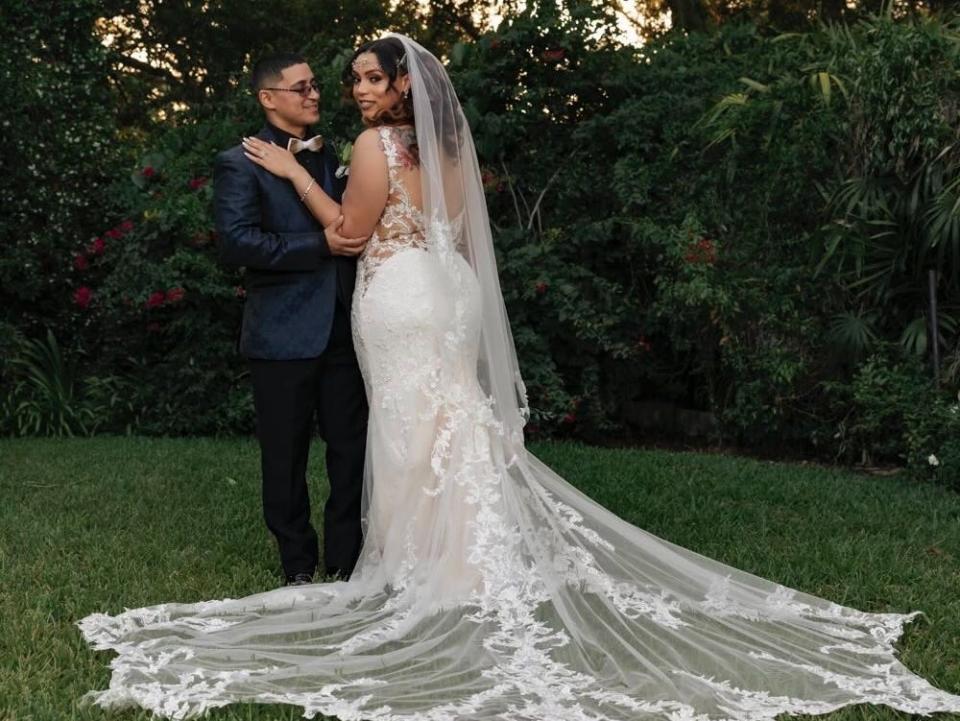 Lilly Mendoza standing next to a man with a blue suit and a golden bowtie with her back turned so the trail of her wedding dress is spread out.