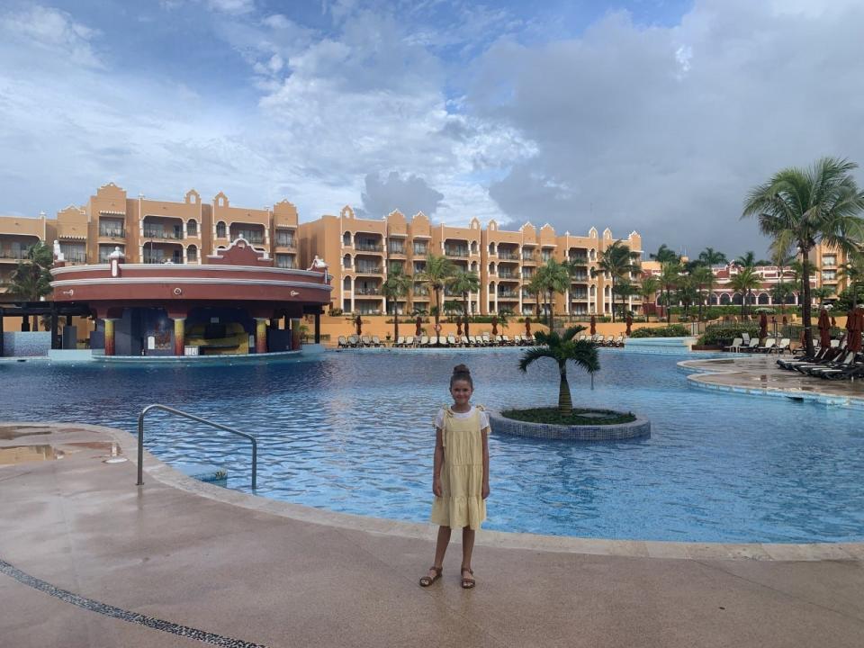 A girl posing in front of a large hotel pool with a palm tree in the middle of it.