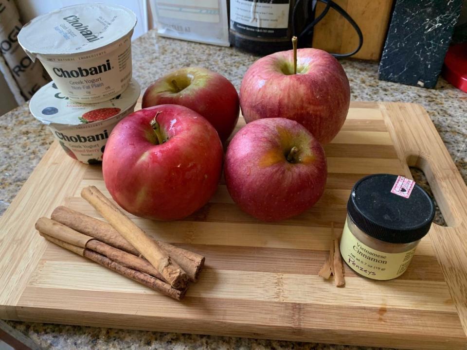 a cutting board with four apples, cinnamon sticks, a jar of ground cinnamon, and greek yogurt