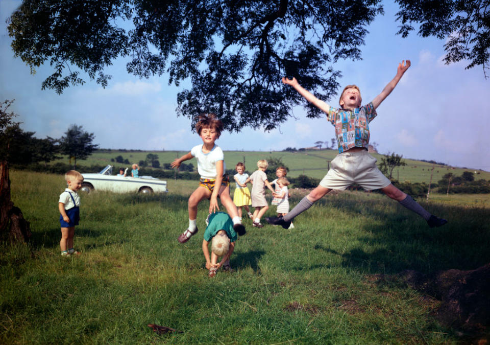 Children playing and jumping in a field with a classic car parked nearby, surrounded by trees and rolling hills