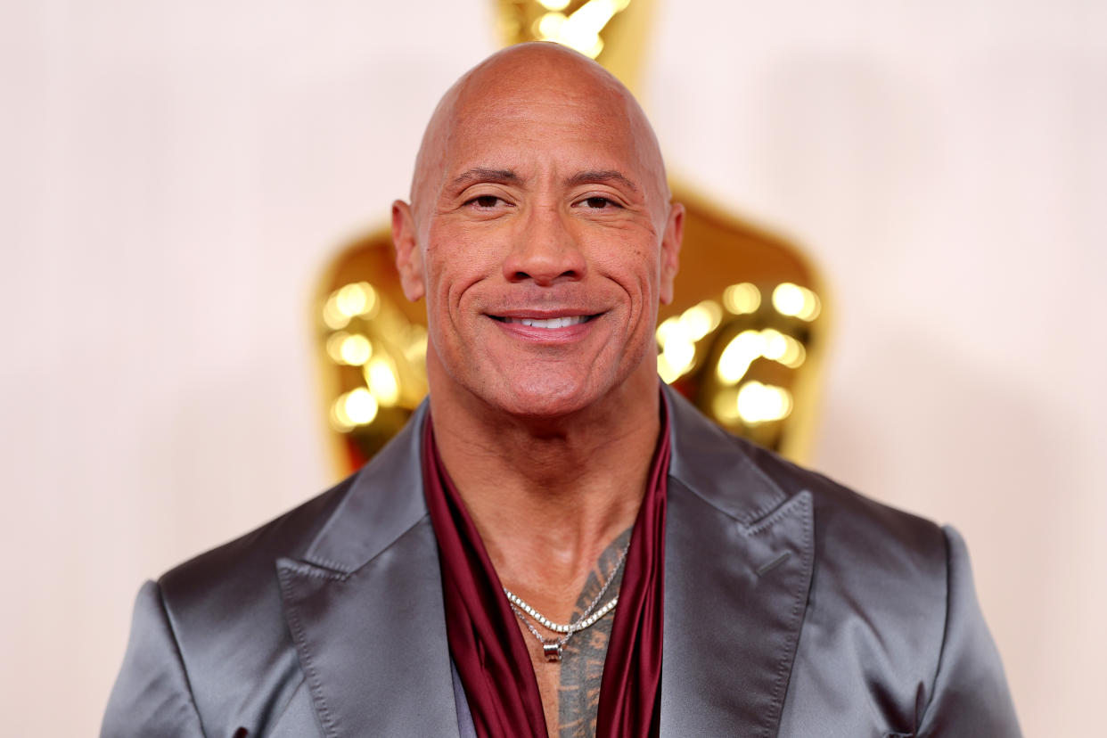 Dwayne Johnson smiles for the camera at the Academy Awards. A large gold Oscar statue can be seen behind him.