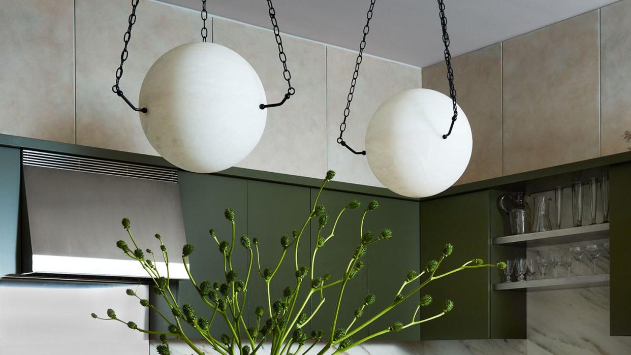 a kitchen has an island with a marble surface and dark olive green cabinets, two alabaster globe pendants suspended by chains, marble backsplashes, stainless oven, sink with open shelving and glasses above