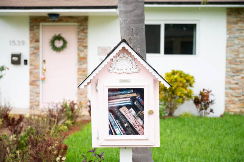 little pink house-shaped free library full of books posted on tree trunk in front of white and pink house. Green lawn and some bushes around