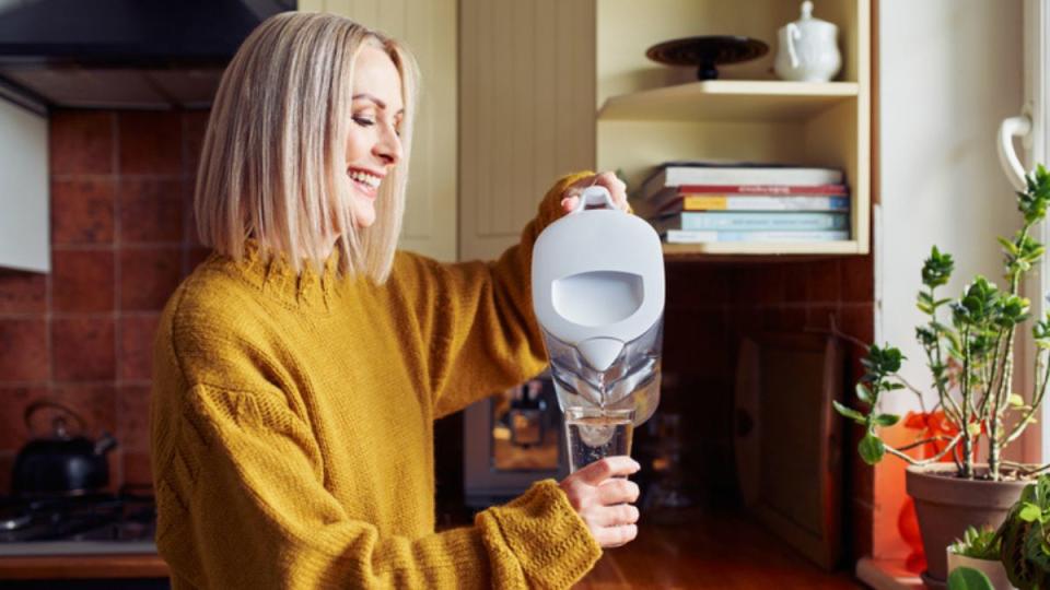smiling mature woman pouring filtered water into a glass in the kitchen to hydrate to prevent waking up dizzy