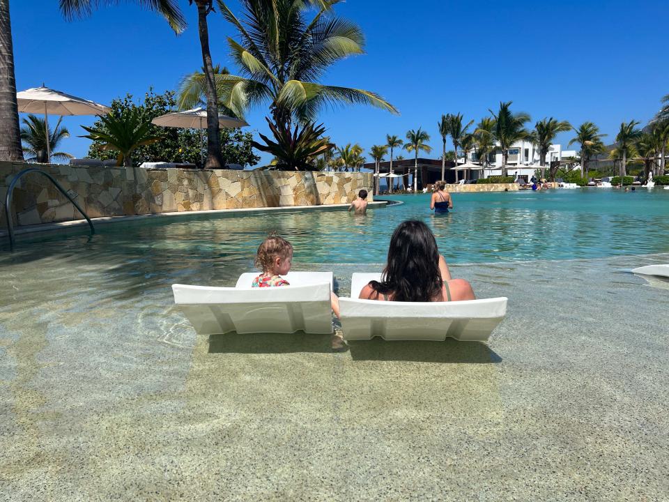 A woman and child sitting on chairs in the shallow end of a pool as seen from behind.