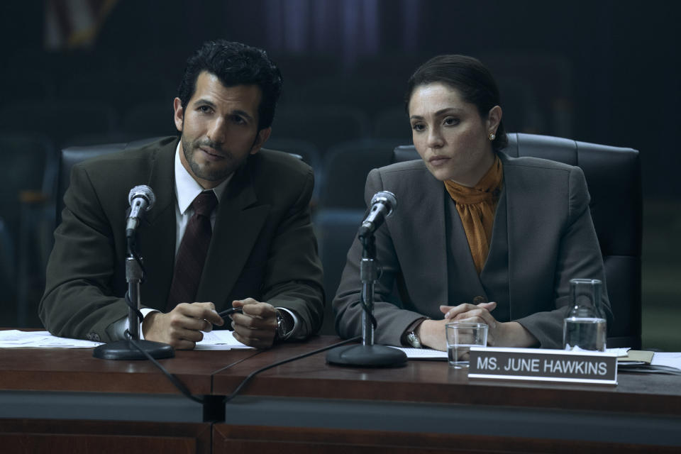 a man and a woman sit at a desk in front of microphones, with a sign in front of the woman that reads, ms. june hawkins