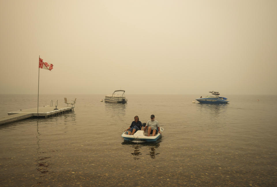 Smoke fills the air as Pat Manzuik and his wife Trevor use a paddleboat to get to shore after being given a boat ride by good samaritan Christy Dewalt, back right, back to their home they were evacuated from due to the Lower East Adams Lake wildfire, in Scotch Creek, Canada, Sunday, Aug. 20, 2023. (Darryl Dyck/The Canadian Press via AP)