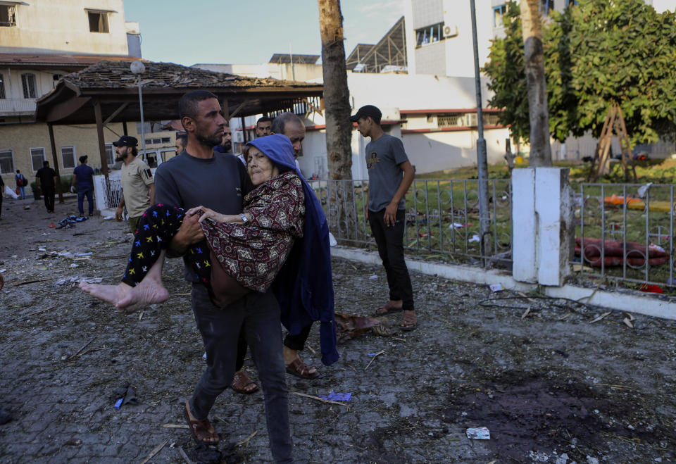 A Palestinian man carries an elderly woman past the site of a deadly explosion at al-Ahli hospital, in Gaza City, Wednesday, Oct. 18, 2023. The Hamas-run Health Ministry says an Israeli airstrike caused the explosion that killed hundreds at al-Ahli, but the Israeli military says it was a misfired Palestinian rocket. (AP Photo/Abed Khaled)