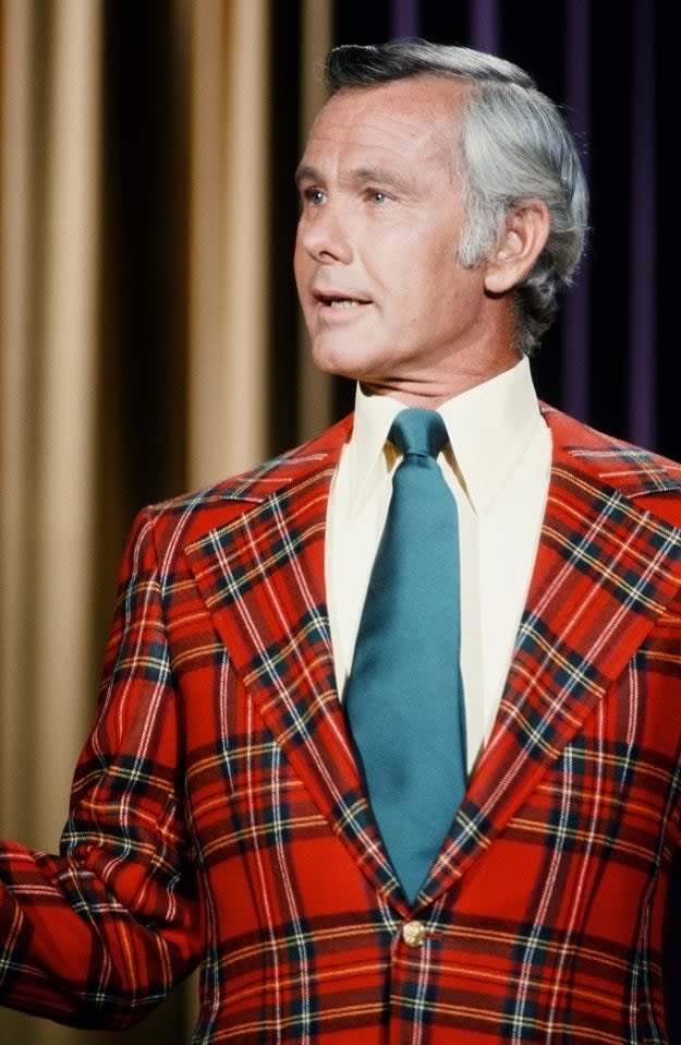 Johnny Carson wearing a plaid suit jacket with a green tie and a yellow shirt, speaking in front of curtain backgrounds