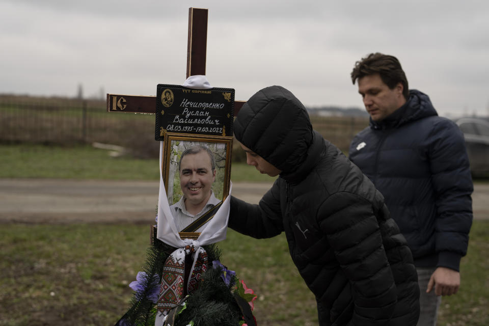 Yura Nechyporenko, 15, kisses the pictures of his father Ruslan Nechyporenko in front of his uncle Andriy Nechyporenko, at the cemetery in Bucha, on the outskirts of Kyiv, Ukraine, on Thursday, April 21, 2022. The teen survived an execution attempt by Russian soldiers while his father was killed, and now his family seeks justice. (AP Photo/Petros Giannakouris)