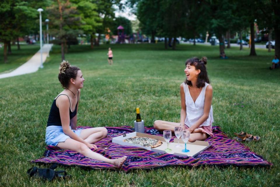 Two women in a park together with a bottle of wine and three glasses, one woman is laughing very hard.