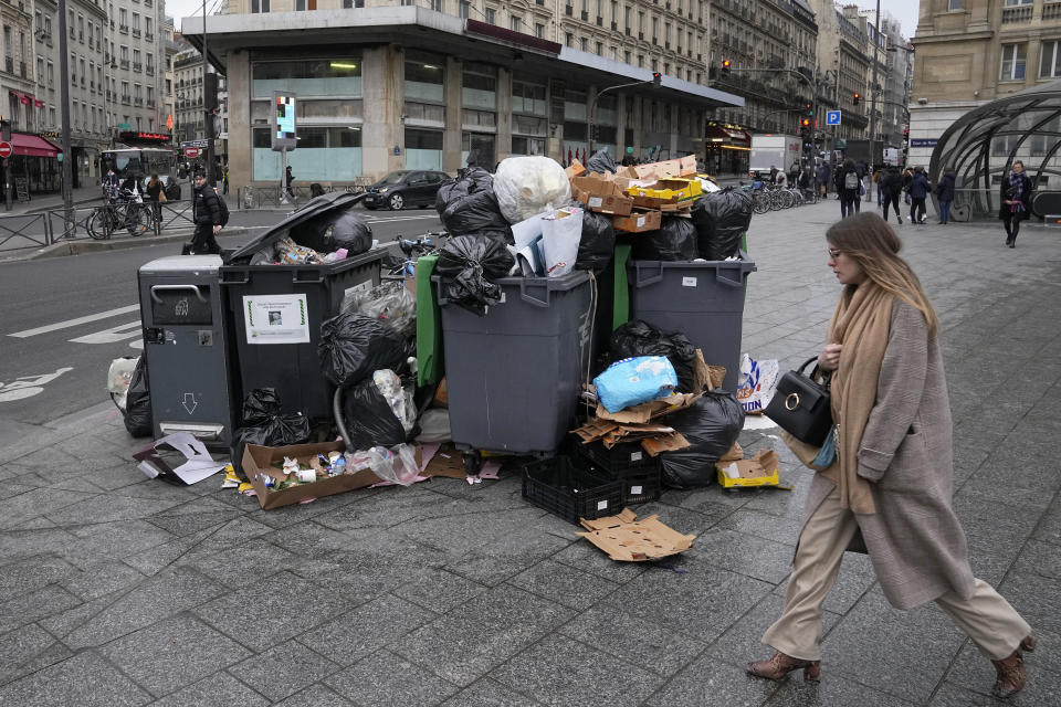 A woman walks past a pile of garbage cans Tuesday, March 7, 2023 in Paris. Garbage collectors, utility workers and train drivers are among people walking off the job Tuesday across France to broadcast their anger at a bill raising the retirement age to 64, which unions see as a broader threat to the French social model. (AP Photo/Michel Euler)