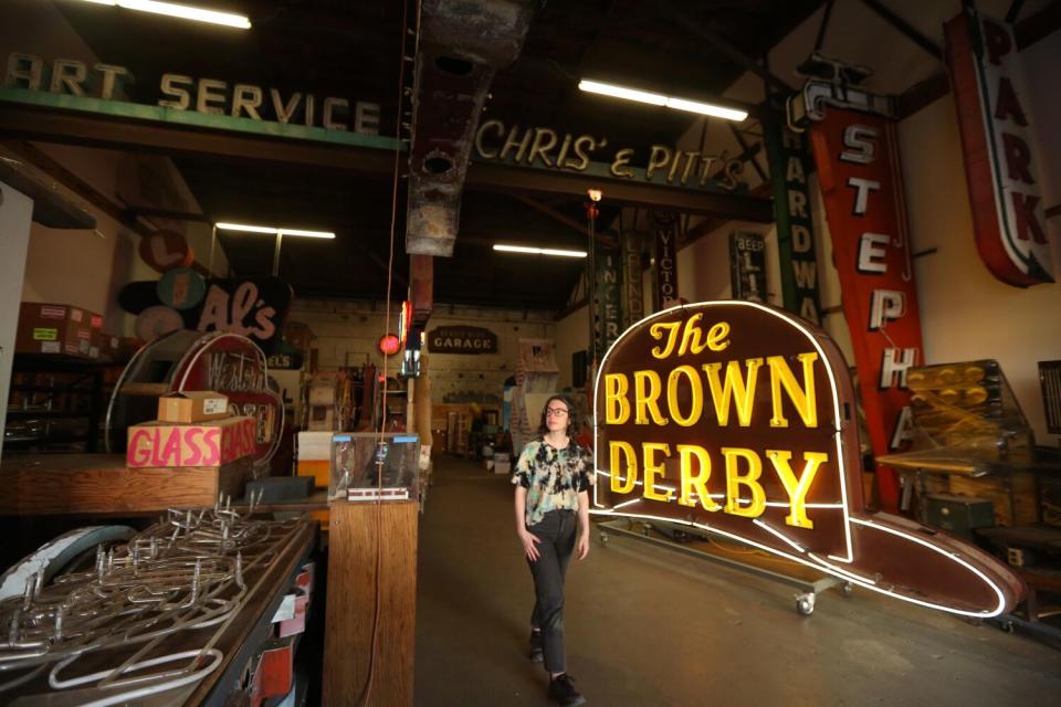 a woman stands in front of a "The Brown Derby" neon sign