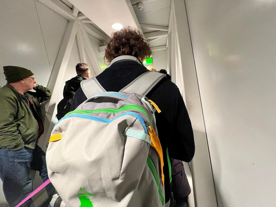 Bored passengers stand in a small corridor at Stansted Airport waiting to board a Ryanair plane. In the foreground a man has a very large backpack.