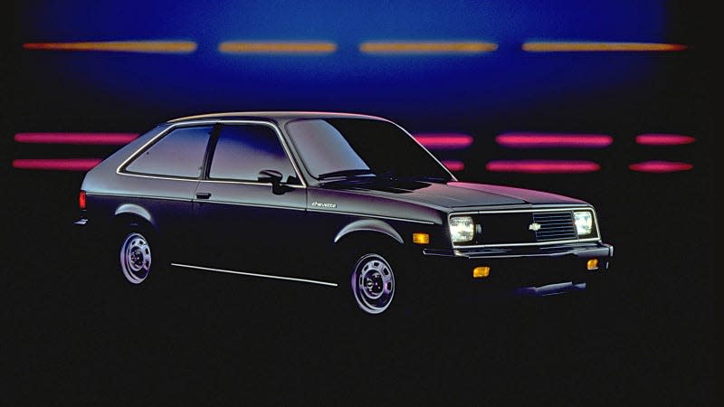 A black Chevette seen from the front quarter in front of a background with yellow and red lights.