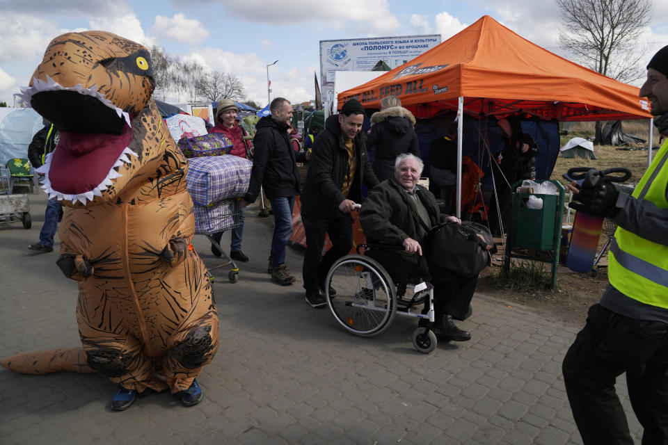 A refugee in a wheelchair smiles as a volunteer dressed in a dinosaur suit stands at the border crossing in Medyka, southeastern Poland, Monday, April 11, 2022. (AP Photo/Sergei Grits)