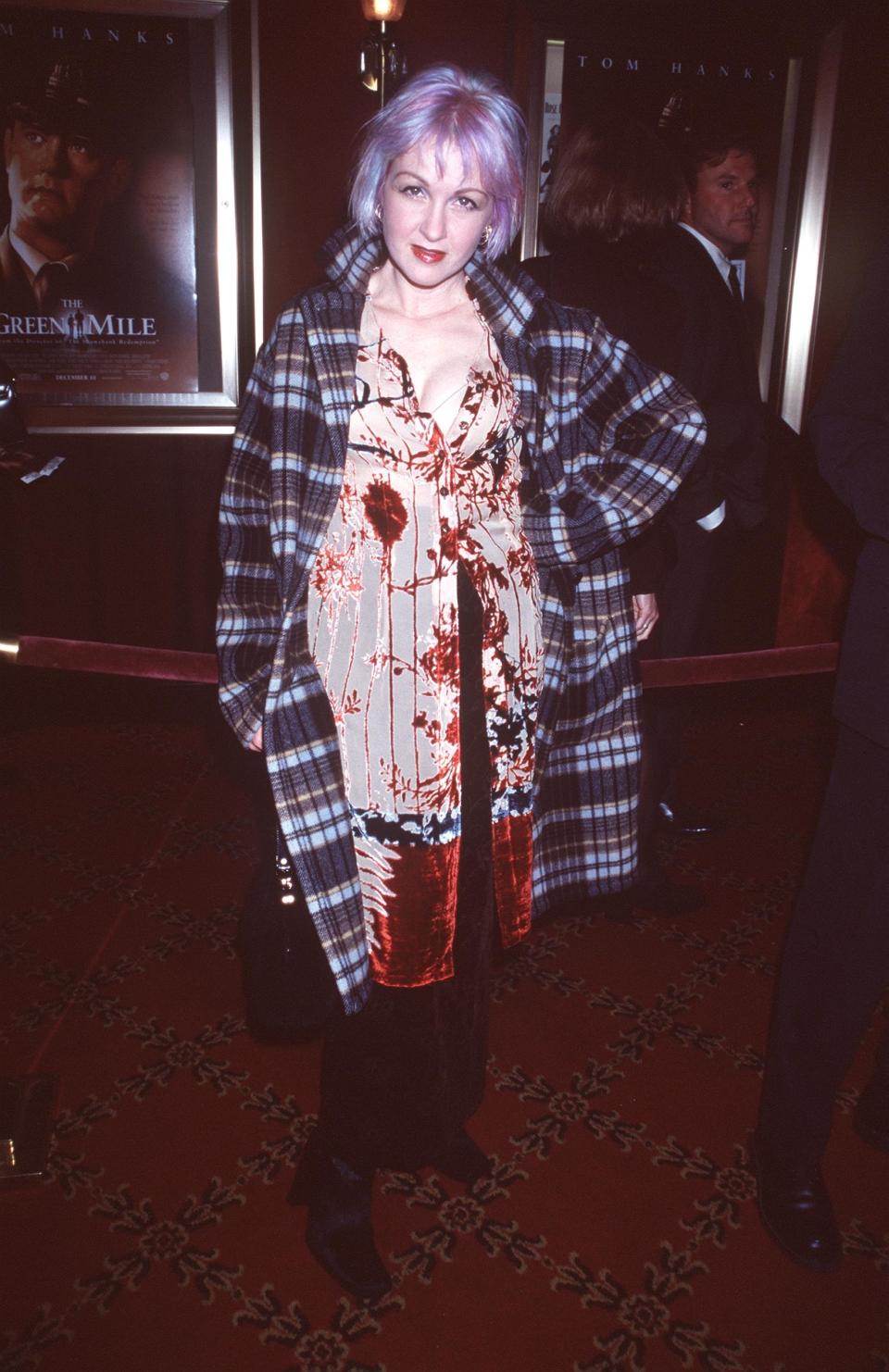 Person on red carpet in plaid coat over patterned dress, standing near a movie poster