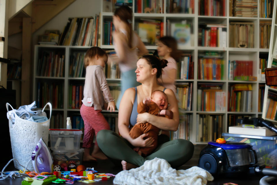 A woman cradles a baby amidst a cluttered living room with toys and laundry. Three other children play and move around her