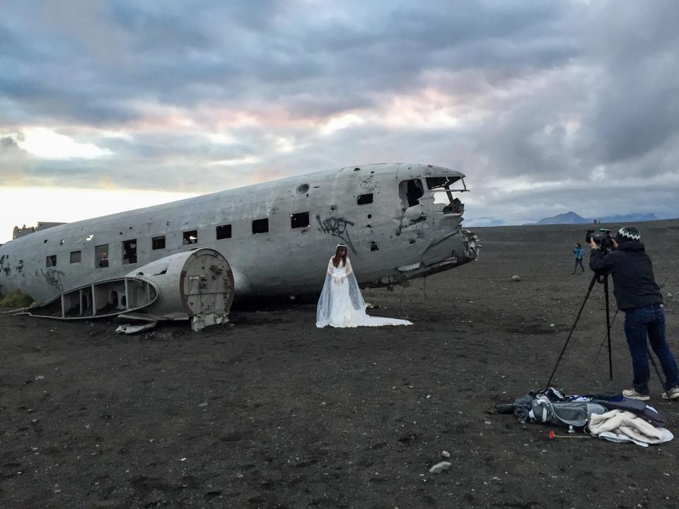 A woman in a wedding dress posing next to the Sólheimasandur Beach plane wreck as someone takes a photo using a tripod nearby