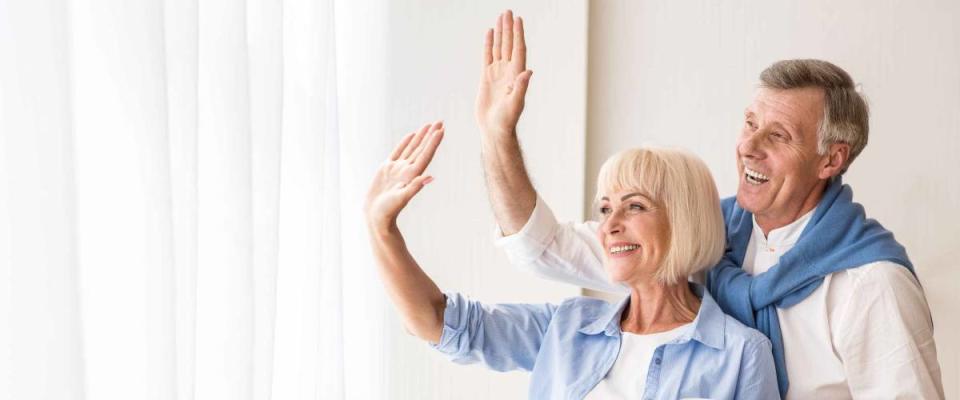 Senior couple waving hands near window, saying goodbye to grandchildren, free space