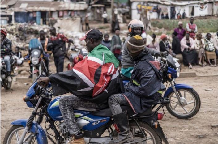 A man on a motorcycle taxi with a Kenyan flag draped over him.