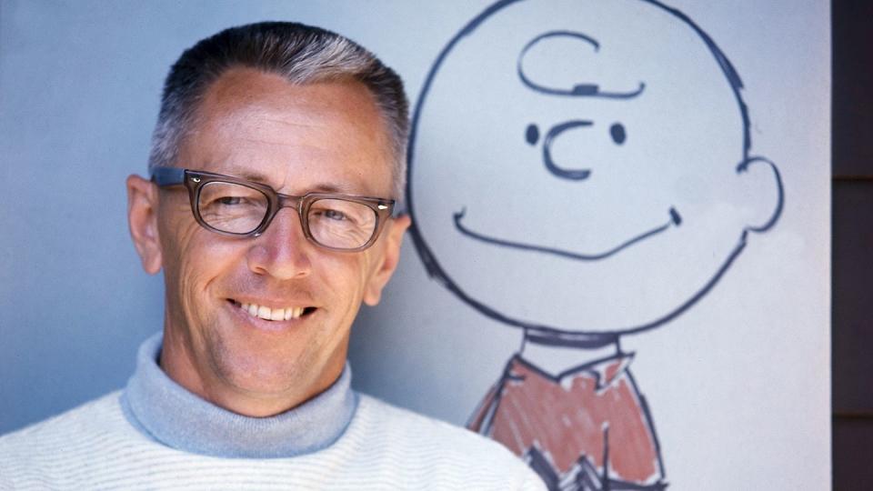 charles schulz, wearing a white and gray turtleneck and glasses, smiles while standing in front of a drawing of charlie brown