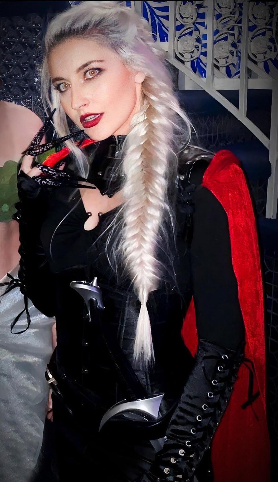 A woman with a long, blond braid holds up her hand, which has long nails. She wears a red cape.