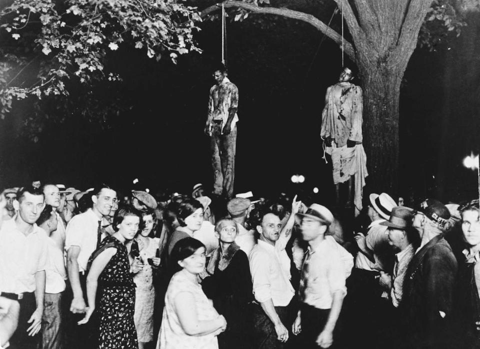 A black and white photograph of a crowd under a tree where two bloodied men were hanged.