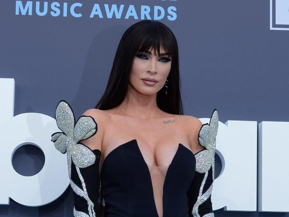 Megan Fox stands on a carpet at the Billboard Music Awards. She wears a black dress with a plunging neckline and silver flowers on the sleeves