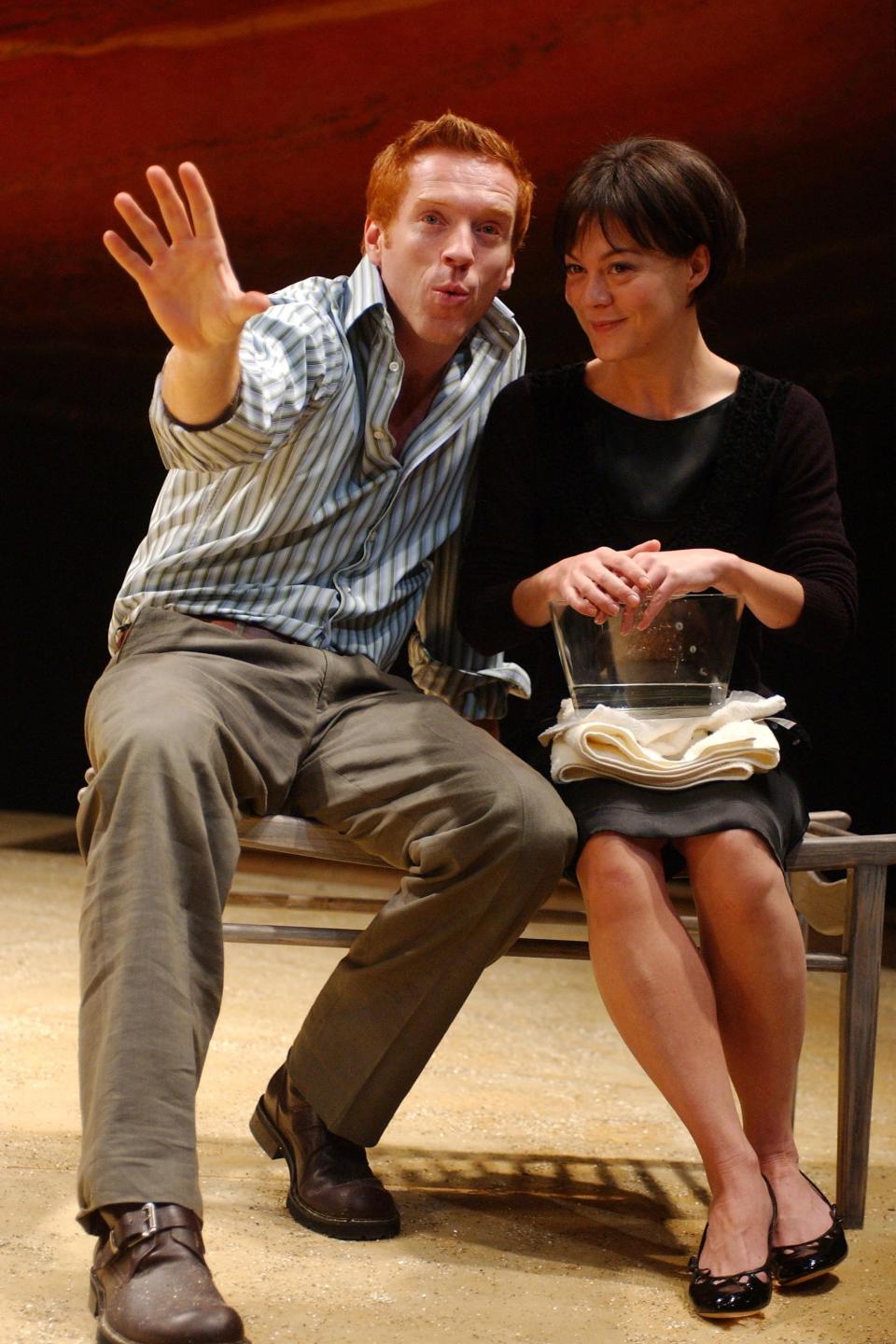 Damian Lewis, in a casual striped shirt and pants, gestures animatedly while sitting beside Helen McCrory, in a simple black dress, holding a glass bowl