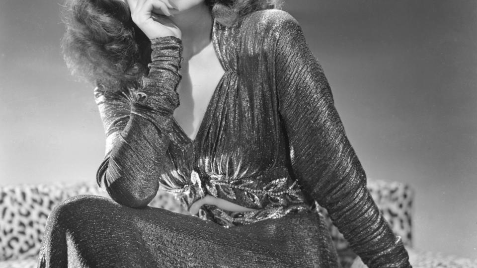 rita hayworth looks off into the distance with a sad face, she wears a shimmery long sleeve dress and sits on a cheetah print armchair