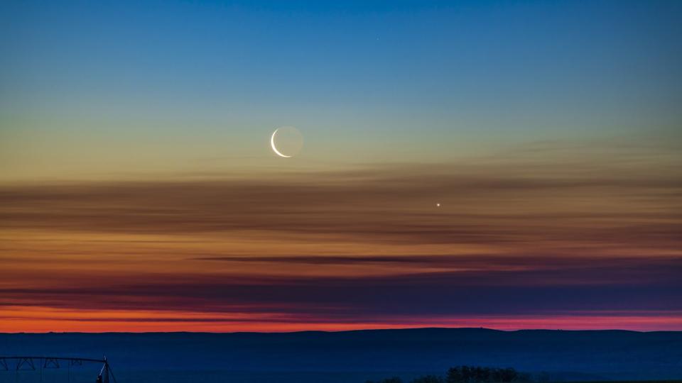 The waning crescent moon conjuncts Venus as it rises low in the northeastern dawn sky over southern Alberta, Canada. Earthlight is visible on the dark side of the moon. The sky displays a spectacular gradient of colors from orange at the horizon to blues at the top of the spectrum