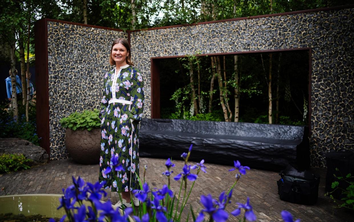 A young woman in a floral dress stands in front of a wall of flints and a bench, with birch trees and irises