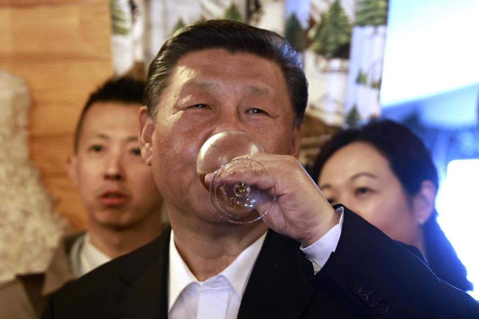 A dark-haired man in a dark jacket and white shirt drinks from a glass