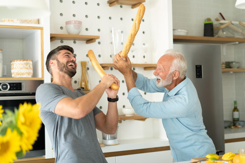 A man and his senior father play fighting with baguettes in a kitchen