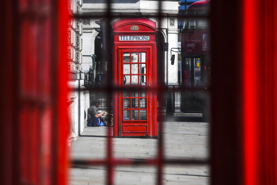 A homeless man sits on the ground, seen through a red phone box window, during lockdown to protect against the Coronavirus outbreak, in London, Tuesday, April 14, 2020. The new coronavirus causes mild or moderate symptoms for most people, but for some, especially older adults and people with existing health problems, it can cause more severe illness or death.(AP Photo/Alberto Pezzali)