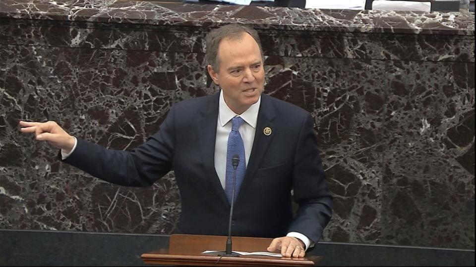 Adam Schiff gesturing in front of a dark marble wall as he speaks into a mic