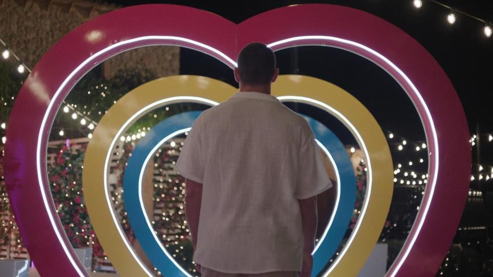 Joey Essex entering the Love Island villa. Joey is pictured from the back as he walks through a multi-coloured heart-shaped arch. He wears a white short-sleeved shirt and is pictured outside the villa at night, lit by festoon lighting. 