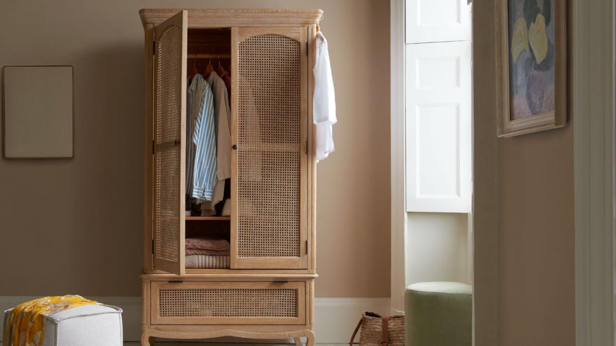  The Margot free standing wardrobe  with a partially open door full of lcothes, beside a small white square pouffe stool 