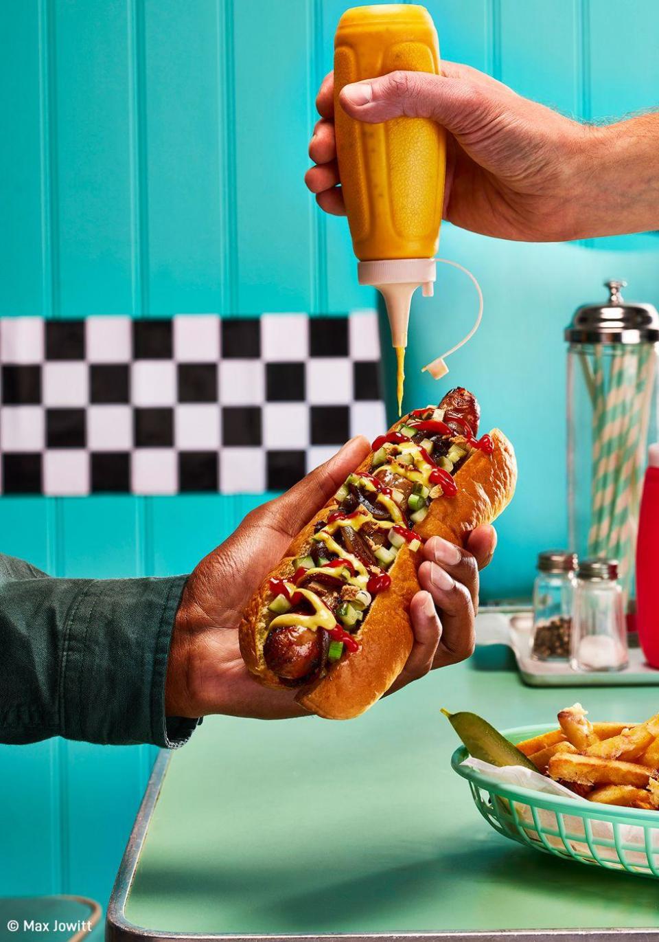 A hot dog, held in one hand, while another squeezes mustard on top