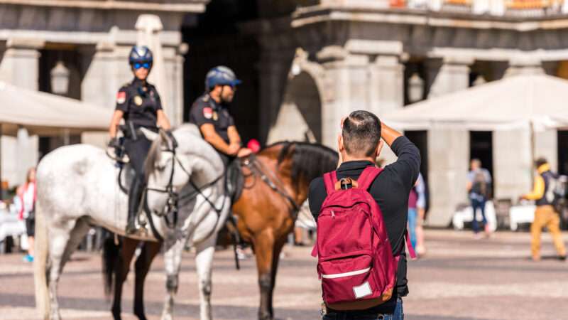 A photographer with a red backpack, in the foreground, takes pictures of police officers on horseback, in the background.