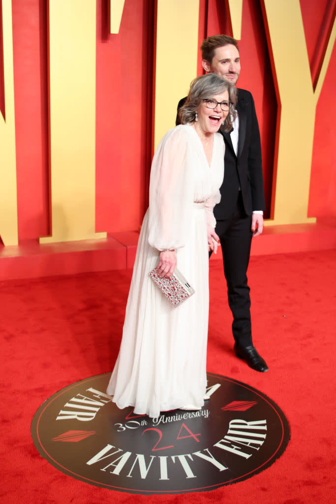 Two people on red carpet, woman in white v-neck dress and man in suit, both smiling