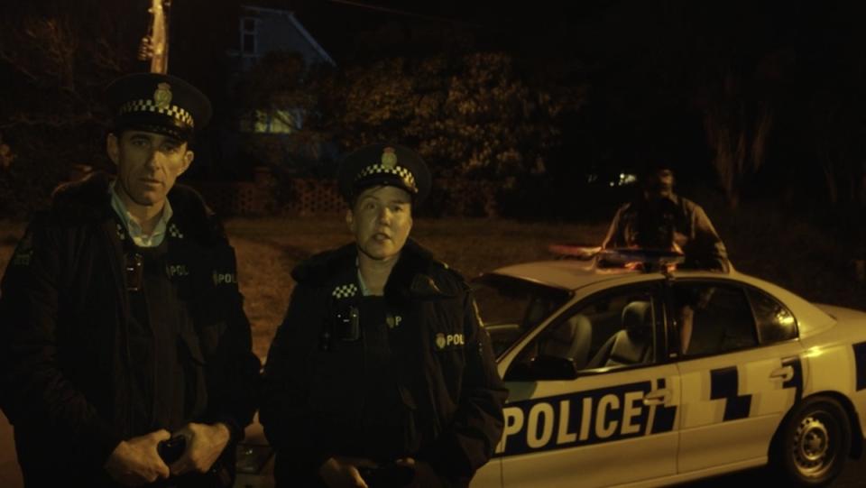 Two cops standing in front of their car on the street outside a house as a ghost stands up in the middle of it