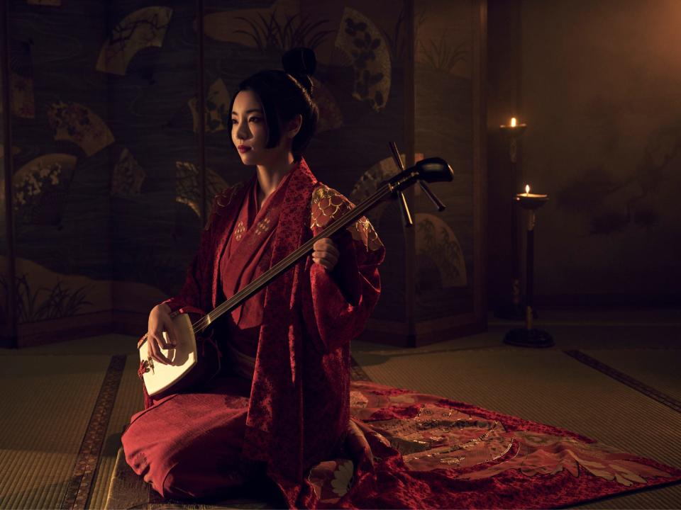 yuka kouri as kiku, ayoung woman wearing ornate red robes, her hair styled eleganty, and playing a shamisen, a lute with three strings and a square body