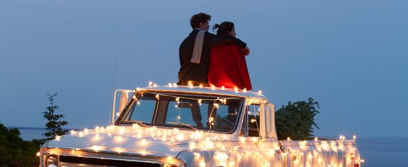 A teen couple sitting on the top of a pickup truck festooned with lights