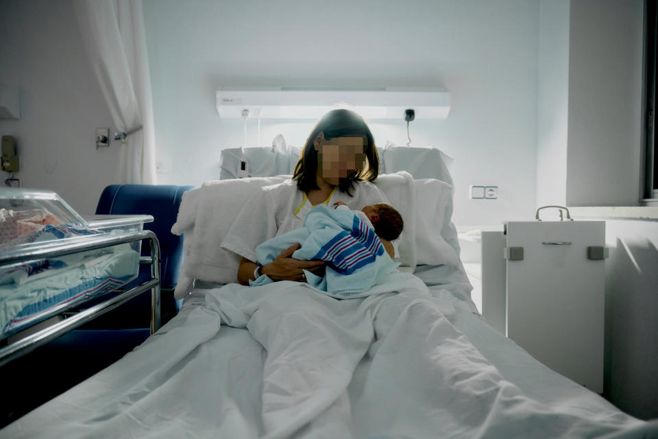 A person holding a newborn in a hospital bed, with a crib and medical equipment nearby. Identity protected with pixelation