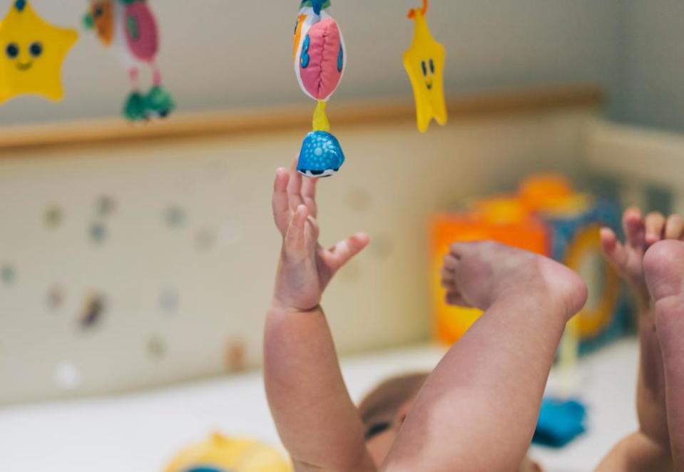 A baby in a cot reaching up to a colourful mobile hanging above her.