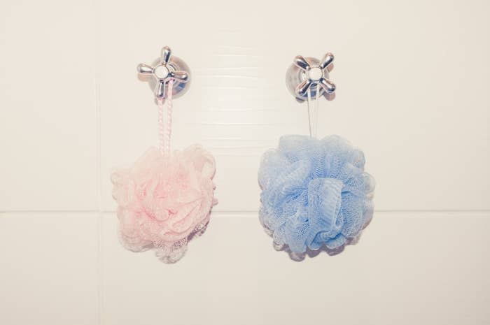 Two bath loofahs, one pink and one blue, hang on silver wall hooks in a bathroom