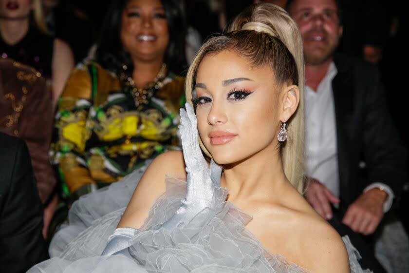 Ariana Grande wears a poufy gown and leans to her left while touching her cheek